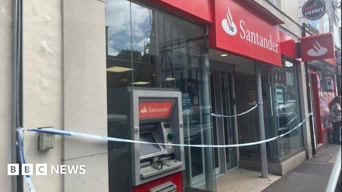 The front of a bank branch with red and white signage including the Santander logo and name. The whole frontage is festooned with blue and white police tape. A cash machine is visible but not obviously damaged from the front.