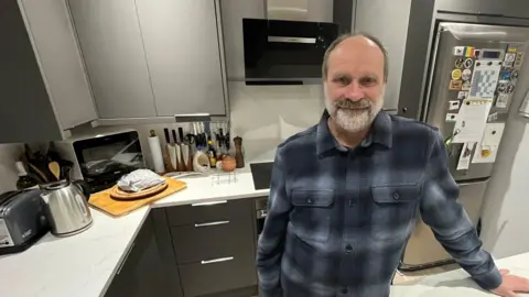 Stephen Gledhill standing in his kitchen. He is wearing a dark blue and white plaid shirt and has a grey speckled beard . Behind him the kitchen is modern with dark doors and drawers and white surfaces