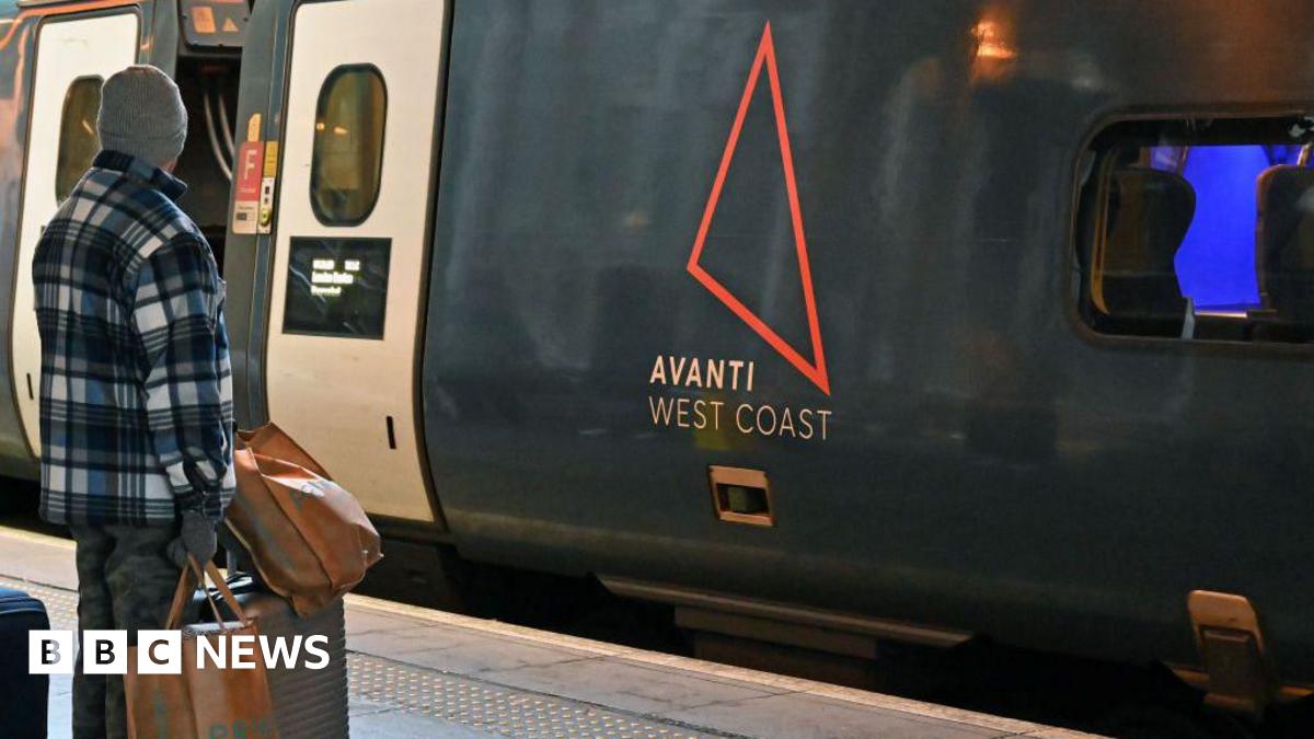 A man in a checked shirt, woolly hat and gloves, stands on a rail station platform. He has his back to the camera and is facing an Avanti West Coast train.