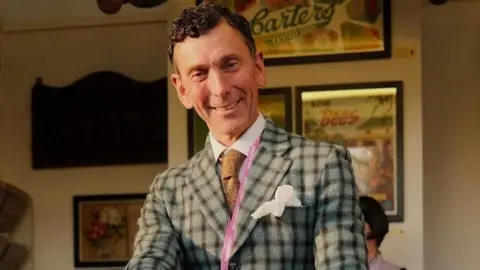 Social media A photo of Matthew Upham looking at the camera. He is wearing a grey checked jacket, a light brown tie and a pink lanyard