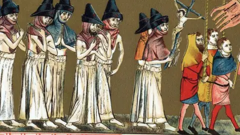 Getty Eight people dressed in white robes and pointed hats walk in bare feet in a procession behind a group of three men in colourful clothes. The men carry flags.