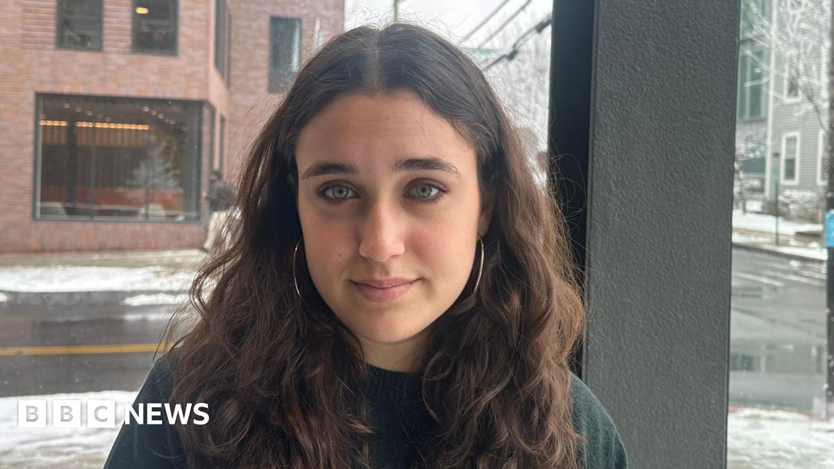 Mia Tretta sits in front of a window in her dorm in Brown University with a street in the background with white snow on the ground. She is wearing a jumper and gold hoop earrings.