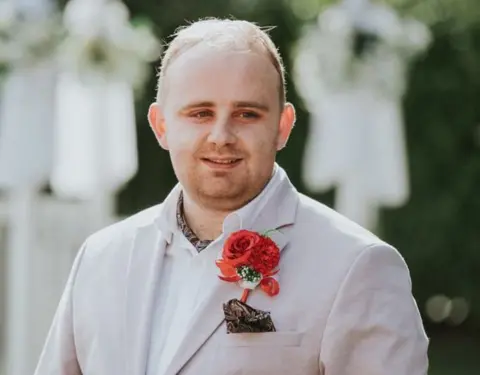 Liam McHale pictured at a wedding, wearing a pale suit with bright red flower in buttonhole. 