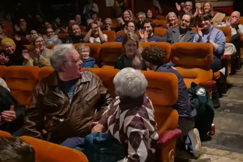 Jacob Molloy/Woolton Cinema Jacob on one knee as Lucy sits in a row of orange upholstered seats in the cinema. They are surrounded by other people who are smiling and clapping