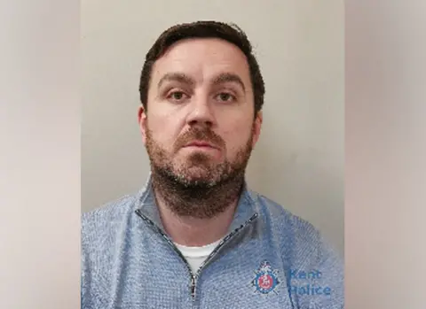 Kent Police A police mugshot of Scott Gunn, a man with brown hair and stubble, wearing a blue jumper.