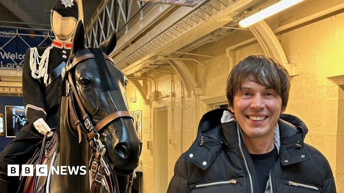 A smiling Professor Brian Cox standing in front of a model of a black horse with a mounted police officer mannequin on it. He is smiling and wearing a black padded coat. The background shows prison cells lit with yellow light