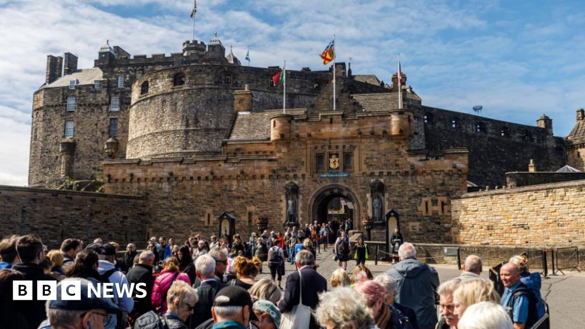 A crowd of people filing into Edinburgh Castle, a large medieval-style castle. Steep stone walls, turrets and towers rise from a flat entrance flanked by smaller walls. Colourful flags flutter against a blue sky.