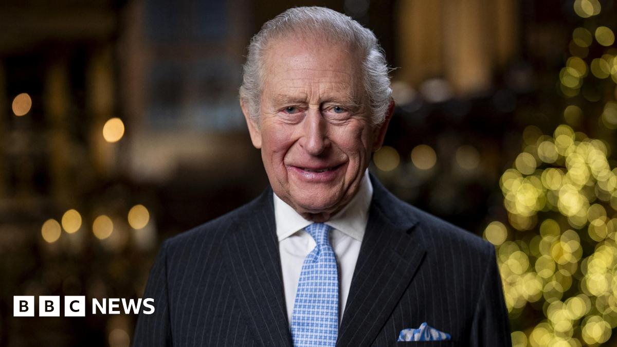 King Charles, wearing a pinstriped suit and pale blue tie and pocket square, smiles at the camera, with the lights from a Christmas tree just seen in the background