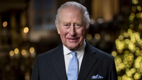 PA Media King Charles, wearing a pinstriped suit and pale blue tie and pocket square, smiles at the camera, with the lights from a Christmas tree just seen in the background 