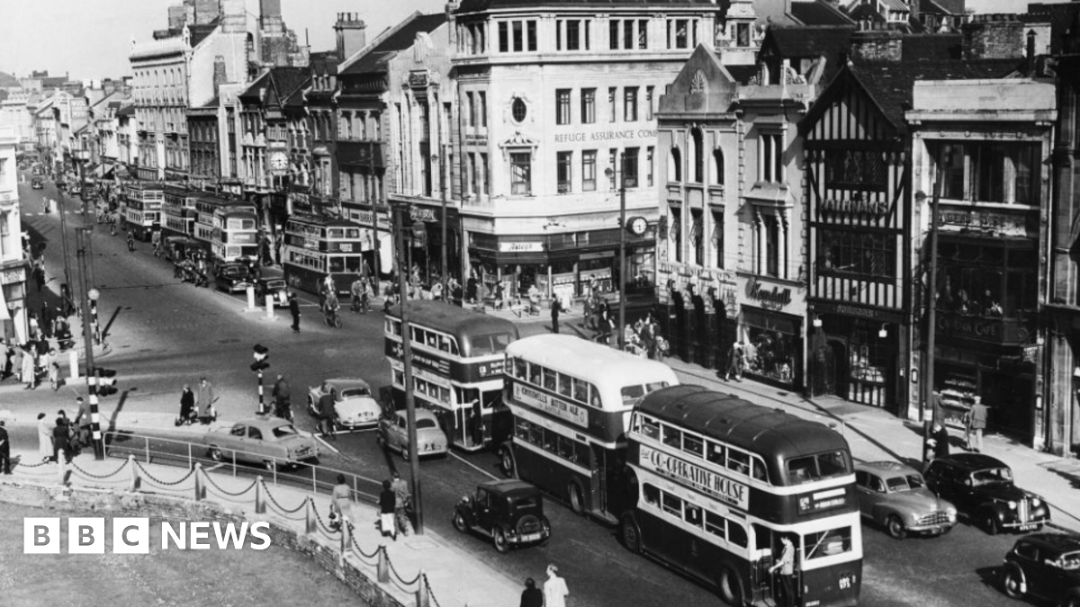 A black and white photo of Cardiff city centre. There are lines of shops on one side, castle green on the other. Through the middle is a road with double decker buses and cars on it and there are lots of people milling about.