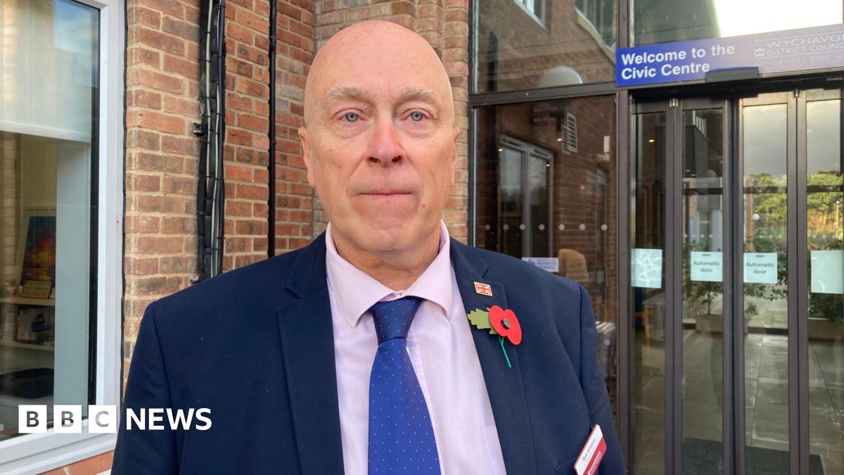 A man wearing a blue shirt, pink shirt and blue spotted tie, stood behind a council building.