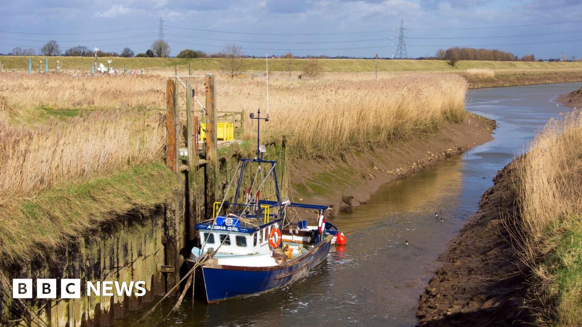 The mouth of the Vernatt’s Drain where it feeds into the River Welland near Spalding, The eastern bank of the Welland can be seen crossing the centre of the image and there is a blue and white boat in the foreground.