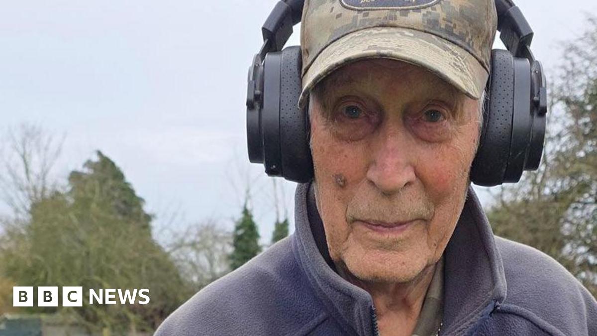 Dave Derby standing outside in a field, wearing a faded camouflage base ball hat, with large black wireless headphones over the top. He is also wearing a grey fleece. Behind him is grace and trees.