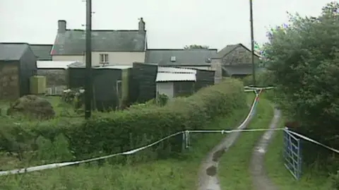 A shot of a farmhouse among bushes and green shrubbery. There is a path to the right side with a metal gate that is open.