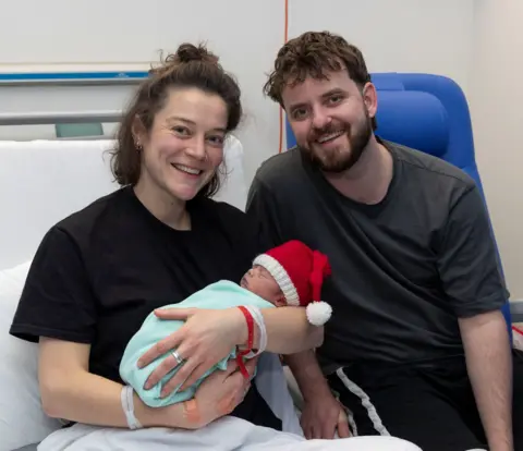Alan Simpson A smiling woman wearing a black t-shirt and holding a new born baby wearing a santa hat. Next to her is a smiling man with a dark t-shirt. 