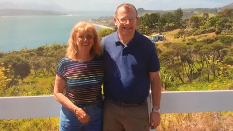 Claire Corkery Sue and Tom a smiling older couple on holiday with a lush green landscape and bay behind them
