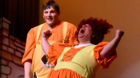 Camphill Milton Keynes Two people in orange and yellow costumes stand on stage. One is shouting and waving their arms. The other is laughing.