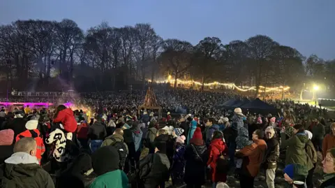 Jacob Tomlinson/BBC A large crowd of people spanning across a park. There are several trees in the background with hanging lights illuminating the early evening darkness. 