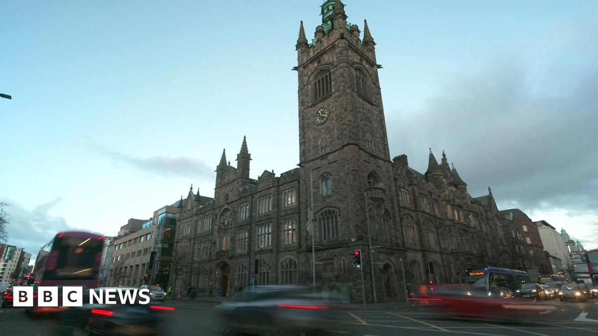 A large church building in Belfast city centre shows, stained glass windows and a bell tower, bearing a gold and black clock. Behind the building is a gloomy, cloudy sky. Traffic is blurred as it passes the camera.