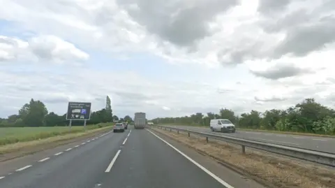 Google A general view of the A1 near Stibbington. Vehicles travel on each side of the carriageway on a cloudy day. There is a brown road sign detailing a truck stop nearby.