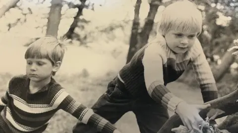 Angela Bushby Two boys, both wearing horizontal striped jumpers, are next to a tree. One of the boys steadies it while the other, looking determined, is about to scale it. 