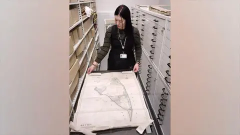 East Riding of Yorkshire Council A woman wearing a long-sleeved dark green top and black dress with long brown hair looking down at a large vintage map that is spread out on a table. There are draws next to her and cardboard boxes on the shelves.