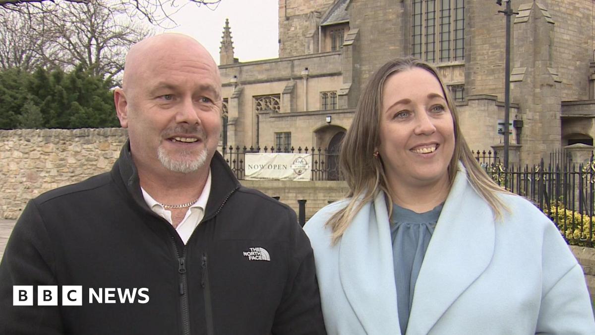 Simon and Tanya Brown standing side by side outside a large sandstone building with decorated windows and spires. Mr Brown is bold and has a grey goatee. He is wearing a black zip-up fleece over a whites shirt and he is wearing a silver chain around his neck. Mrs Brown has shoulder-length light brown hair and is wearing a baby blue coat over a grey-blue blouse. They are speaking to a reporter who is interviewing them out of shot.