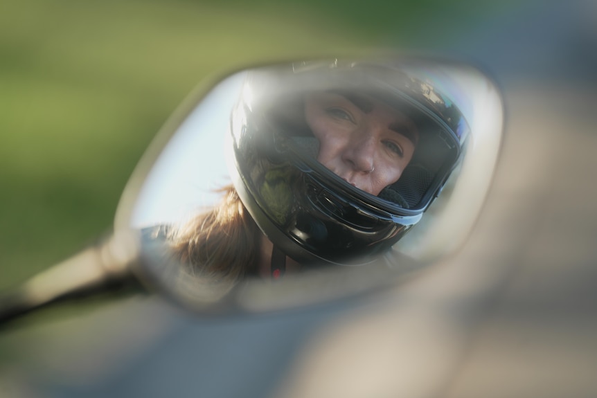The reflection of a woman in a motorbike helmet in a side mirror of a bike.