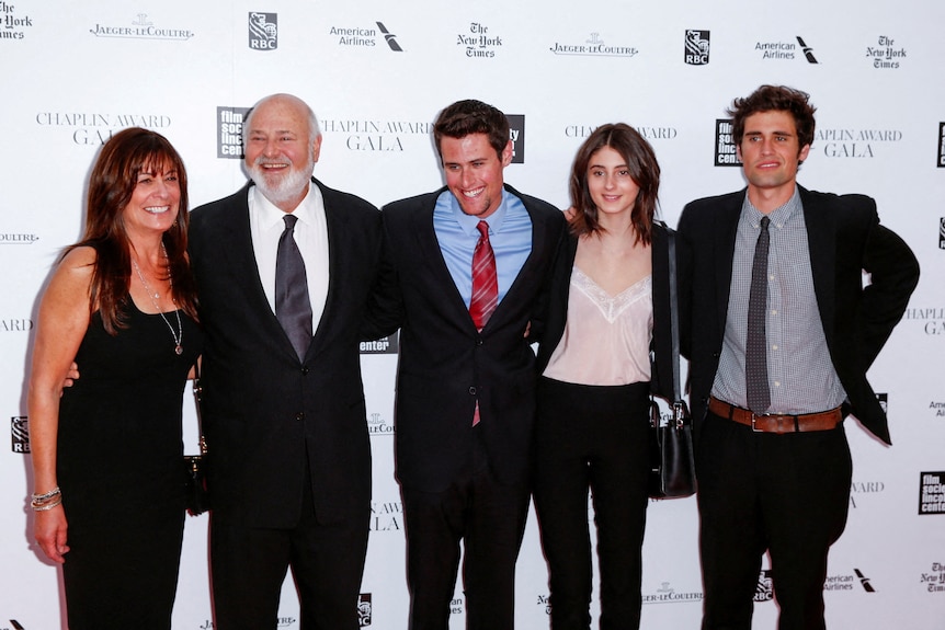 A family embrace each other arm-in-arm and smile in front of a white wall on a red carpet