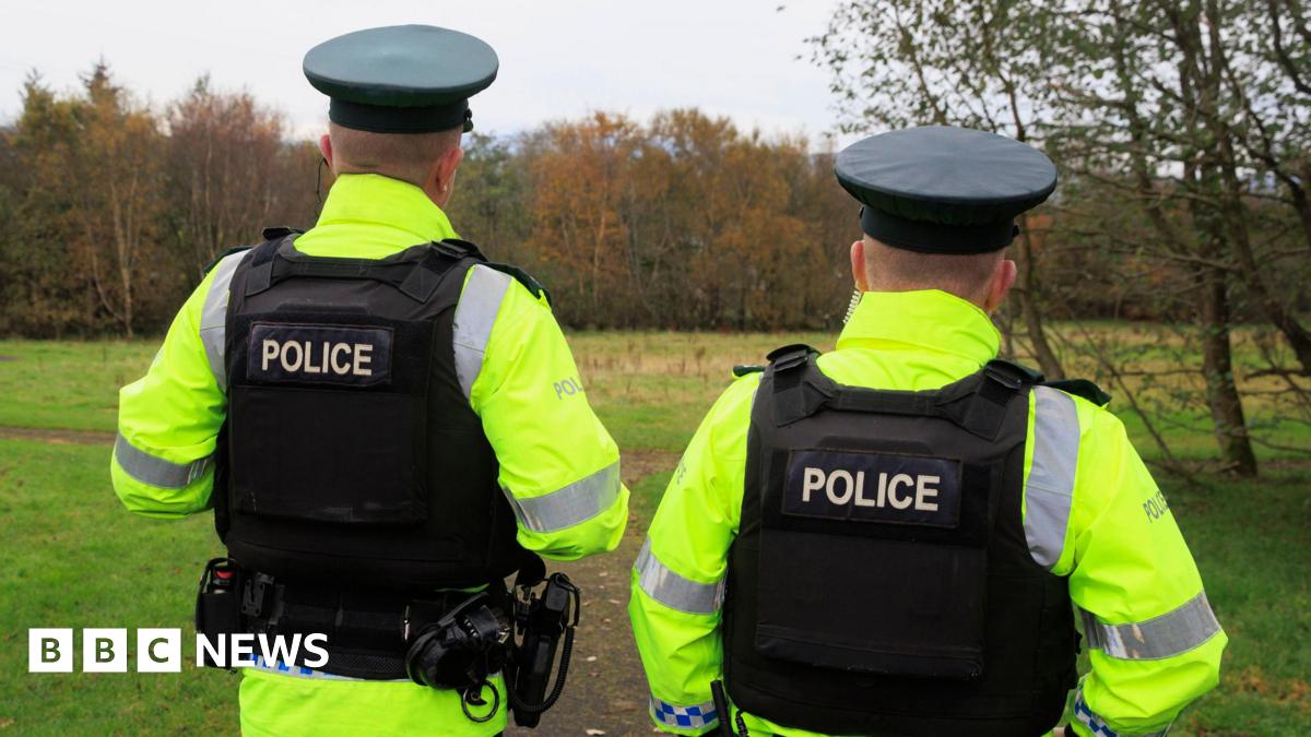The back of two police officers. They are standing in a park with a row of trees in front of them. Both officers are wearing black caps, hi-vis coats and black vests reading "police" on the back.