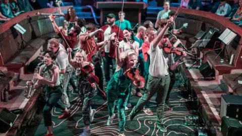 Edinburgh's Hogmanay The band, playing an array of instruments are leaping out of the picture. They are earing green trousers and white tops.