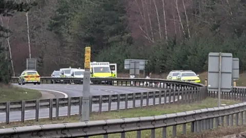 Jack MacLean/BBC The scene of the incident and police response as seen from a distance. At least five police response vehicles are on the road, along with an ambulance and two other vehicles which cannot be identified. Forensics teams are surveying the carriageway.