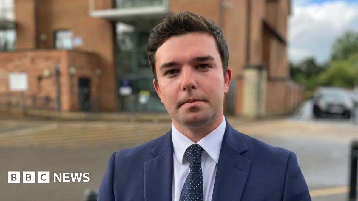 Alex Wagner has brown hair and is wearing a dark navy suit jacket, white shirt and navy tie with white polka dots. He is standing in a cark park which has the brown-brick council building in the background
