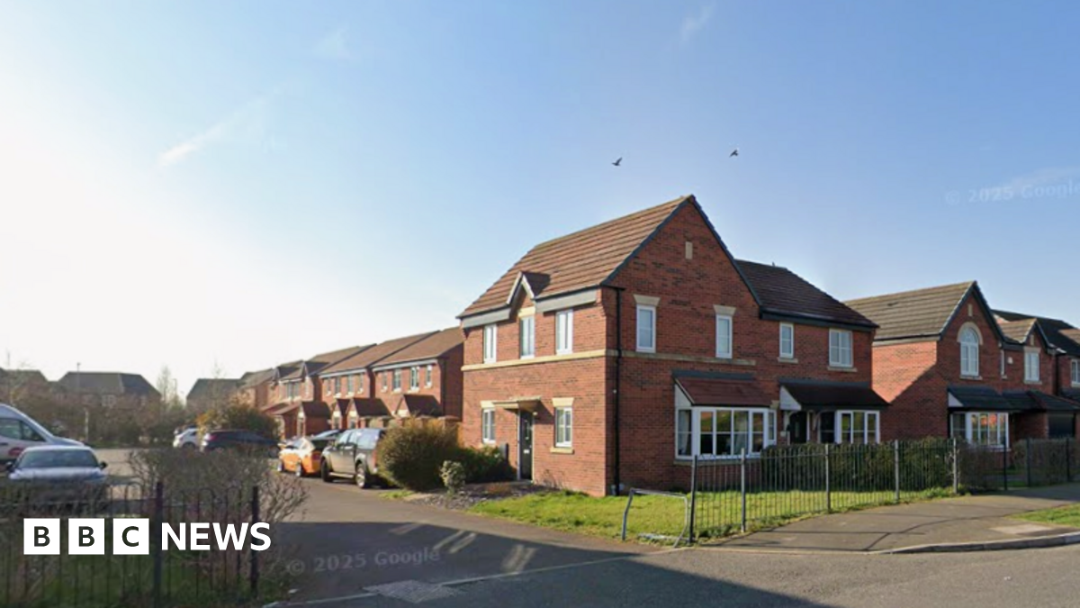 The opening of a road with houses and cars along it. They are set against a blue sky, and there is railing along a pavement in the foreground of the image.