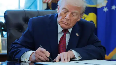 Getty Images A middle-aged man in a blue business suit, white shirt and red tie signs a document at a table. 