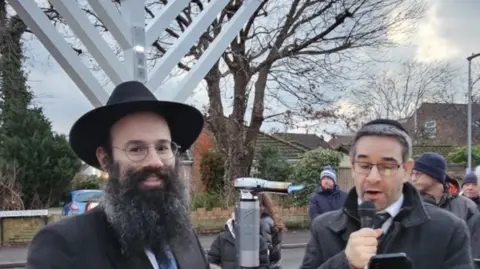 Chabad Lubavitch Bricket Wood Rabbi Tunk smiles at the camera while standing in front of the white Menorah, which is not yet lit. He wears a black hat and black suit jacket with a blue tie. He has a long full beard and wears glasses. The other Rabbi is clean shaven and wears a skull cap and grey winter coat. He is speaking into a microphone while looking at his phone. People are crowded around behind them.