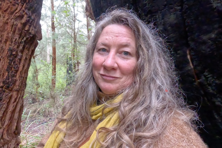 A woman with long grey-brown hair in a forest.