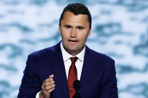 Getty Images Charlie Kirk is standing behind a podium, wearing a dark blue suit with a white dress shirt and a shiny red tie. A black microphone is positioned in front of the podium. The background features a soft, out-of-focus pattern in light blue and white tones, creating a formal event or conference setting. One hand is raised slightly, suggesting he is speaking or gesturing during a presentation.