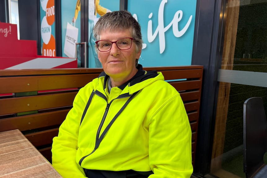 A middle-aged woman in glasses and a high-vis jumper sits at a table outside a cafe.