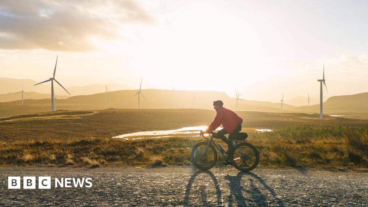 A man in a red jacket rides a bike along a gravel road, past wind turbines as the sun sets in Scotland.