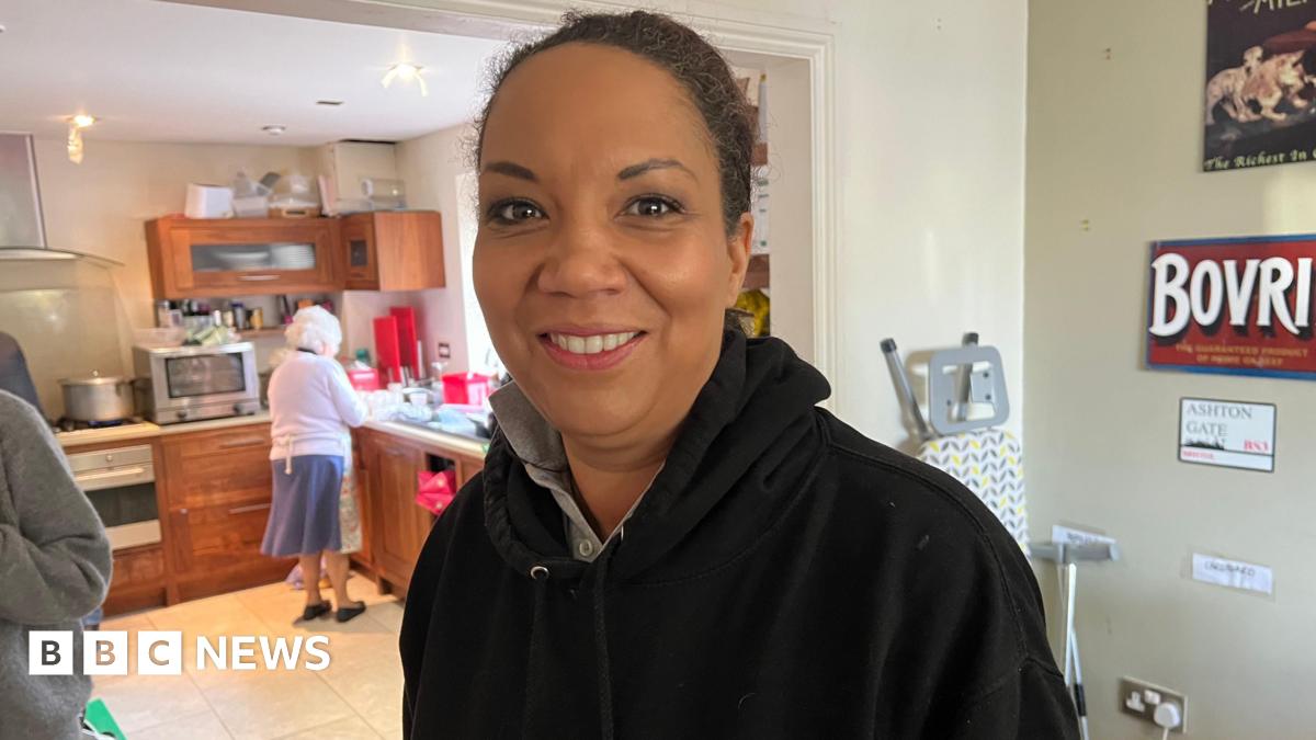 Caz Goddard is smiling, wearing a black hooded jumper while stood near a kitchen. An elderly woman is pictured doing the washing up behind her.