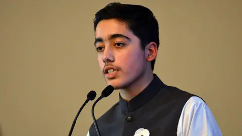 Getty Images A teenager speaks into two microphones as he addresses an audience off-camera. He's standing in front of a plain light brown background and wearing traditional dress - a high-collared waistcoat over a white shirt.