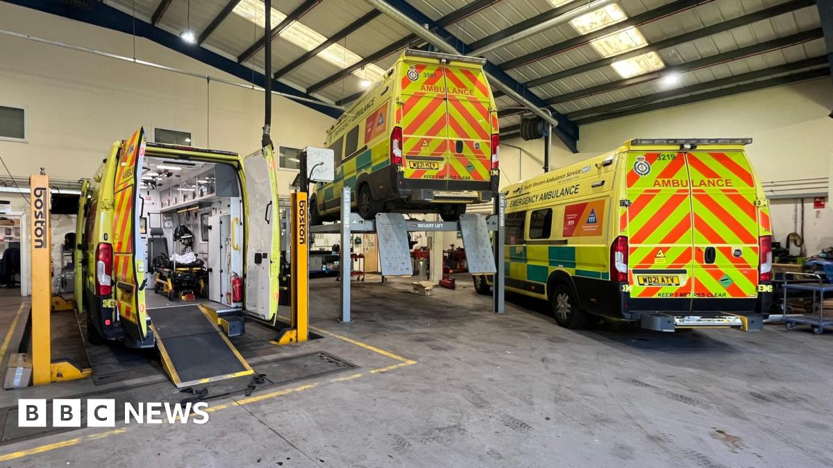 Three ambulances are being worked on inside a depot. Two are on hydraulics, with the middle ambulance lifted into the air. The rear doors are closed on the third ambulance.