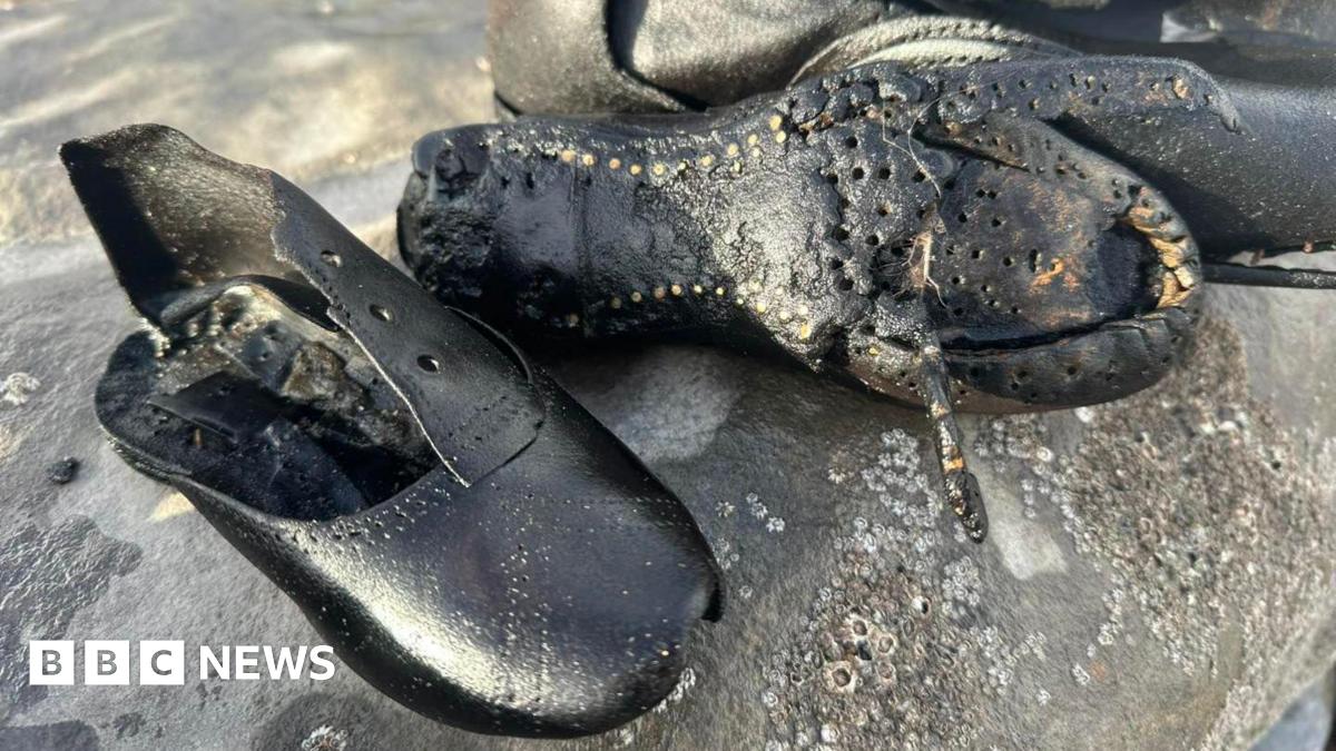 Two black leather boots are on a rock covered in barnacles. The laces are missing. The damaged sole of one of the boots can be seen, along with nails.