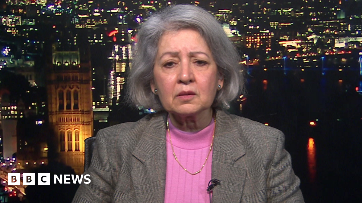Baroness Sheehan is looking at the camera, has brown eyes and grey hair. She is wearing a pink jumper and a grey blazer with a gold necklace.