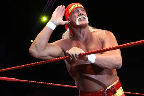 Getty Images Hulk Hogan is inside a wrestling ring, leaning on the red ropes with one arm while raising the other hand to the ear in a listening gesture. He is wearing a bright red bandana, matching red trunks with a yellow belt, and has long, light-coloured hair flowing down the back. The background is dark with spotlights illuminating the scene, creating a dramatic atmosphere typical of a professional wrestling event.
