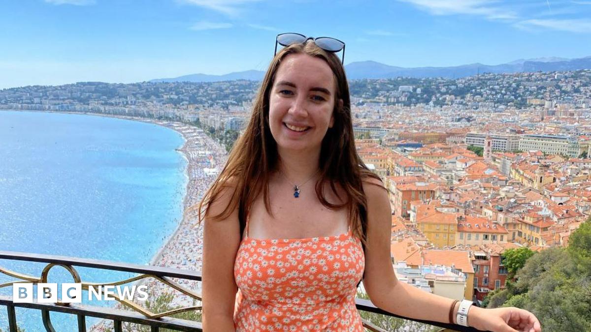 Claire wearing an orange sundress overlooking the coastline of Nice, France. There are orange buildings in the background the sea on the right.
