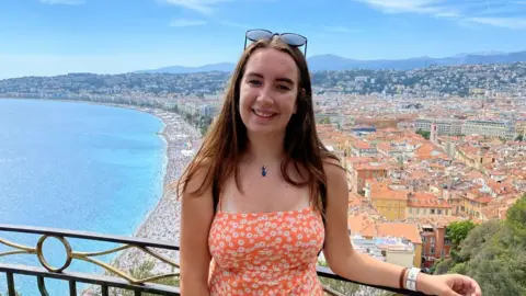 Claire Thomson Claire wearing an orange sundress overlooking the coastline of Nice, France. There are orange buildings in the background the sea on the right.