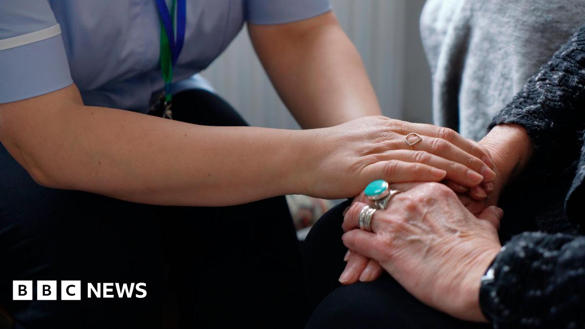 A nurse holds the hand of an elderly lady. They're both sat down.