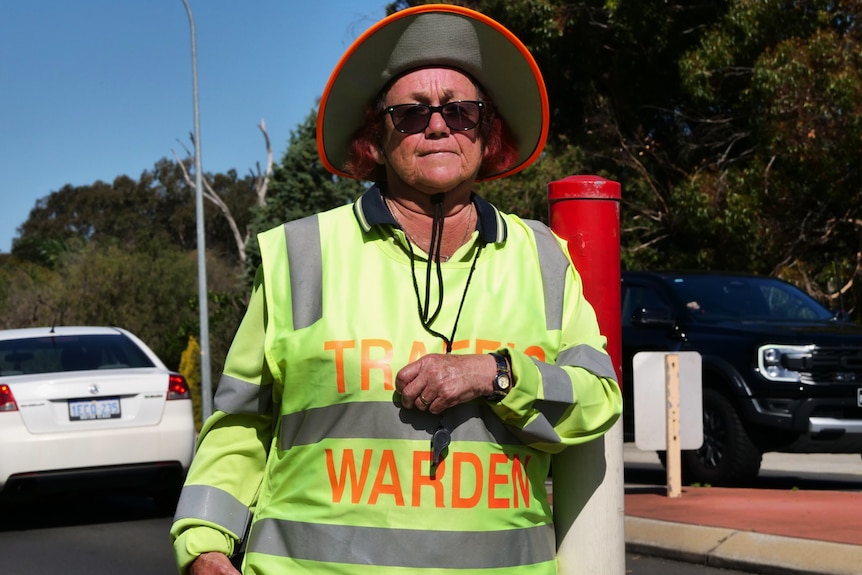 A middle-aged woman in sunglasses, a hat and a high-vis vest that says "Traffic warden" on it.